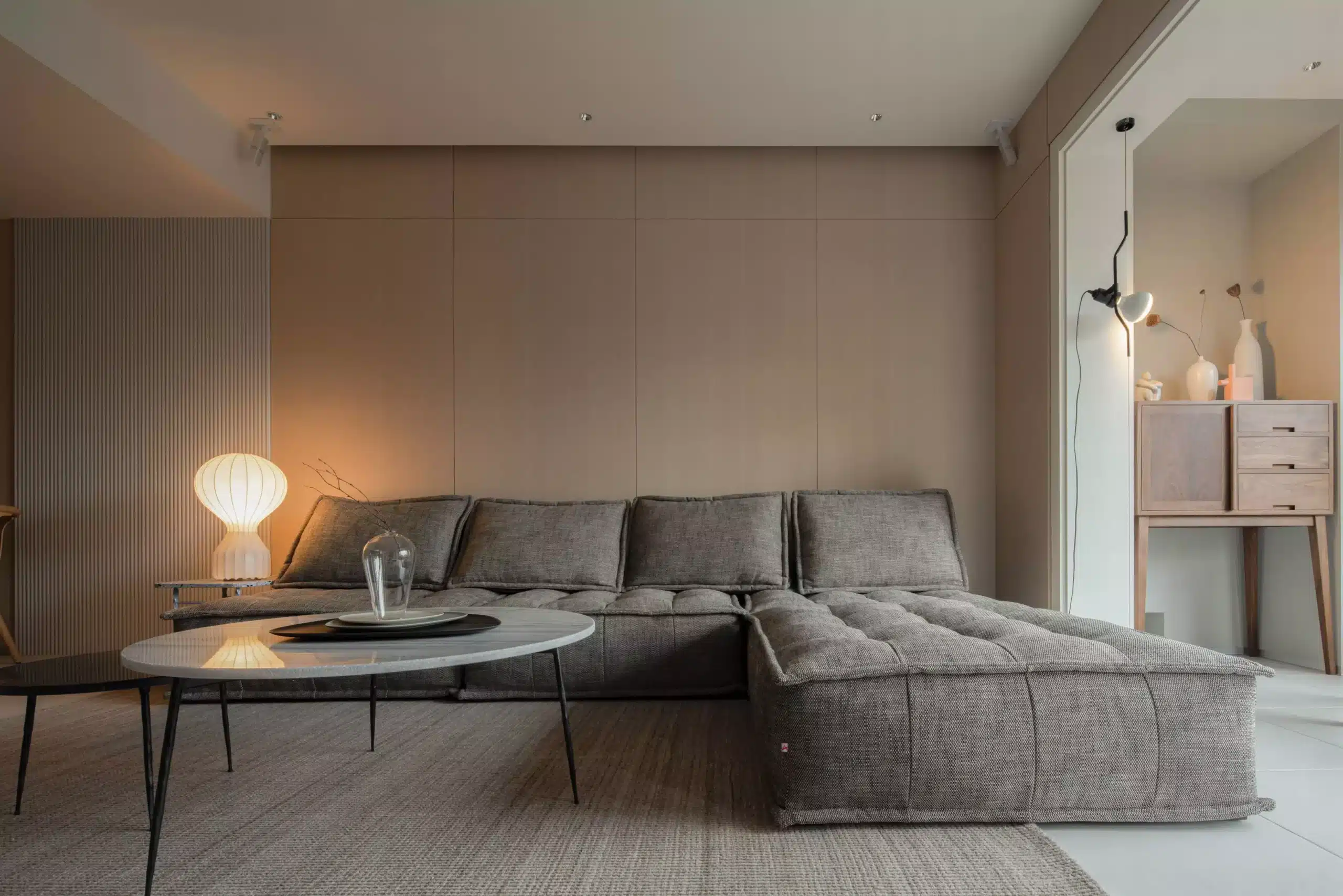 日式侘寂風客廳，配置低矮塊狀灰色沙發與圓形石材茶几，牆面採用溫潤木飾板並搭配褶皺地燈，營造沉穩靜謐感。